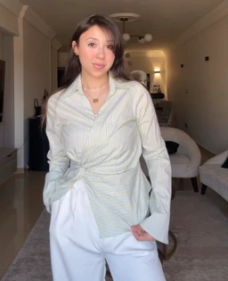 A striped twisted shirt featuring a light base color with subtle vertical stripes. It is styled on a female model, suggesting a unisex-to-women’s fit with a relaxed, oversized vibe and a front twist knot detail at the waist. Visible attributes include a collared button-down shirt with long sleeves, a wrap/twist at the waist, and light, breathable fabric in pale green/blue tones.