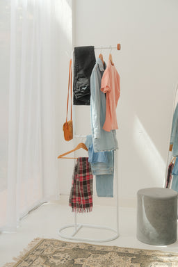 This is a freestanding clothes rack with a slim metal frame displaying a small collection of apparel. The visible items include a mix of lightweight tops and a plaid scarf, suggesting unisex or womenswear; the garments appear in soft pastel and neutral tones. The notable materials look like woven fabrics (knits and cotton-like) with a few solid colors and a red-and-black plaid scarf, all hanging from wooden and plastic hangers.