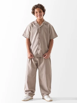 - What the product is: A light, loose-fitting linen pant set consisting of a short-sleeve button-up shirt and wide-leg pants.
- Apparent audience: Boys (young male model) — unisex styling with a relaxed, comfortable fit.
- Notable attributes: Natural linen-like fabric in a beige/stone color; short-sleeve camp collar shirt with front buttons; elastic or relaxed waistband on the wide, baggy pants; white sneakers completing the casual, breathable look.