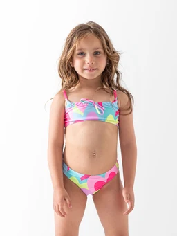 Two-piece swimsuit for young girls. The model is a small child, suggesting a girls’ sizing. The swimsuit features a bright, multicolor tropical/heart pattern with pink, blue, yellow, and green tones. The top is a bandeau style with thin shoulder straps and a small pink bow at the center; the bottoms are mid-rise with moderate coverage. The overall look is playful, summery, and suitable for a girls’ swimwear audience.