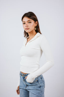 A long-sleeve polo top with a ribbed texture in a solid off-white/cream color. The model appears to be a woman, wearing the top tucked slightly into light-wash denim jeans for a casual, fitted look; the styling is simple and minimal. Notable details include the collared polo neckline and the slim, body-hugging silhouette.