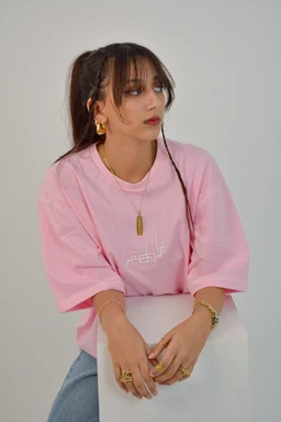 - Product: A pink oversized short-sleeve sweatshirt, worn by a female model. The text on the shirt is subtle and white, with a simple logo/graphic on the chest.
- Audience: Women's/misses (adult female) based on styling, fit, and accessories.
- Notable attributes: Soft pastel pink color, relaxed/boxy silhouette, short puff sleeves, lightweight cotton or cotton-blend material. The styling includes gold jewelry (hoop earrings, rings, necklace) and side-parted fringe hairstyle. No brand visible.
