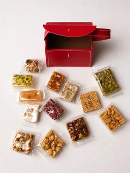 - What it is: A small red jewelry-style box with a flap lid presented open, accompanied by an assortment of small individual packet goodies laid out around it.
- Audience and styling cues: The setup and scale suggest a unisex or gift-oriented product, with a bright red compact box and neatly packaged bite-sized treats arranged for a marketable photo.
- Notable attributes: Bright red box with white stitching, small clear snack packs in various colors and textures (nuts, dried fruits, seeds, crackers), minimalistic white background emphasizing the product. The contents are visible through transparent packaging, highlighting variety and color.