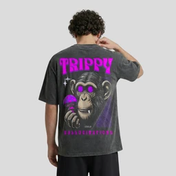 A washed, oversized unisex T-shirt featuring a bold graphic on the back. The model is a man, suggesting a unisex fit with a relaxed, oversized cut. Visible elements include a large pink “TRIPPY” text, a cartoon chimpanzee holding a lighter or mushroom-like object, and purple accents in the graphic, with a dark charcoal fabric and slightly distressed wash. Materials and texture implied: soft cotton jersey with a faded, vintage wash; color palette centered on black/gray with bright pink and purple graphic details.