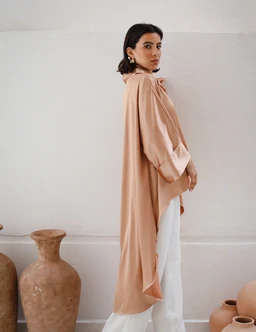 - Description: A long, flowy peach-toned top with a loose, cape-like silhouette and gathered sleeves, styled as a relaxed tunic over white pants. The model is shown in a side profile, standing against a light wall, with minimal accessories and gold hoop earrings.
- Audience and attributes: Appears designed for women; features a soft, drapey fabric with a high, loose neck/turtleneck vibe, oversized sleeves, and an airy, cape-hem silhouette. Notable materials and color: peach/peachy-nude fabric, likely lightweight woven or drapery cotton; creamy white pants for contrast.
- Key style elements visible: oversized, flowing fit; cape-like back/hem; long length; neutral warm color; ruffled or gathered neckline and voluminous sleeves.