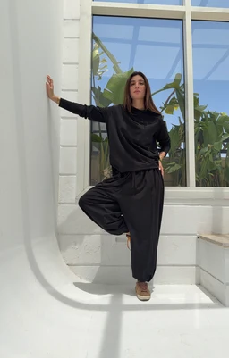This is a black cotton lounge set featuring a long-sleeve top and wide-leg pants. The styling appears to target adults (unisex-leaning but shown on a woman, suggesting women's or unisex casual wear). Notable attributes: solid black color, relaxed fit, soft jersey knit texture, and ankle-to-calf length with a loose, comfortable silhouette. No visible branding; fabric looks breathable cotton.
