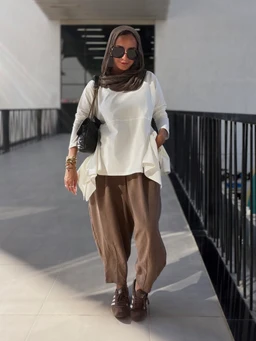 - What the product is: A white tunic-style top with a relaxed silhouette, paired with brown wide-leg cropped pants. The model is accessorized with a brown hijab, dark sunglasses, a small black shoulder bag, and brown lace-up shoes.
- Apparent audience and styling: Appears designed for women; modest, casual-chic styling with loose, layered drape and a comfortable, everyday look.
- Notable attributes: White top with roomy fit and side pockets or draped hem, paired with warm brown earthy-toned pants. Accessories include a brown scarf (hijab), dark sunglasses, and brown shoes; overall color palette is white and brown. The fabric looks lightweight and flowy for the top and soft, drapey for the pants.
