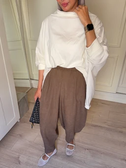 A loose white oversized top with long sleeves tucked partially into high-volume brown balloon pants. The pants are baggy through the hip and thigh with a gathered cuff at the ankle, creating a balloon silhouette, in a warm taupe-brown shade. The look is styled with white flat sandals and a small black handbag, suggesting a casual, minimalist unisex-to-female aesthetic with a soft, breathable fabric appearance.