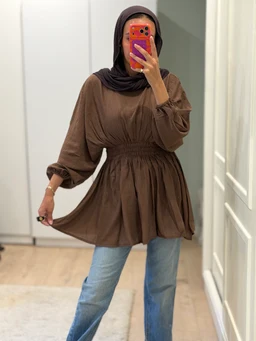 - What the product is: A brown, long-sleeve blouse with a loose, flowy tunic silhouette that cinches at the waist and has a flared, A-line hem. The model pairs it with light-wash jeans and a dark headscarf, taking a mirror selfie.
- Apparent audience: Women (based on styling, hijab-friendly modest fit, and silhouette).
- Notable attributes: Brown fabric, soft and drapey material, voluminous sleeves with elasticized cuffs, elasticized waist for a gathered waistline, and a knee-to-thigh length tunic top with a casual, relaxed style.