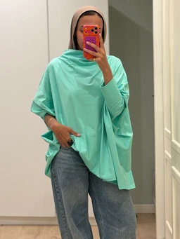 This image shows a loose, oversized mint-green top with a relaxed, poncho-like silhouette and wide sleeves. The model pairs it with light-wash jeans and a beige headband, presenting a casual, unisex-leaning aesthetic. Visible materials look like a soft, smooth cotton or cotton-blend fabric, in a bright mint color with a minimal, drapey style.
