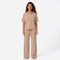A relaxed two-piece loungewear set in a warm beige. The outfit appears designed for adults (unisex-leaning with a loose, casual fit), featuring a short-sleeve polo-style top with a classic collar and a drawstring elastic-waist pant; both pieces are made from a soft, knit fabric with a matte, tone-on-tone finish. Notable attributes include the oversized, drapey silhouette, breathable light-brown colour, and comfortable, easy-wear style.