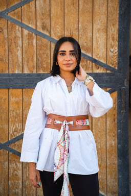 A canvas belt is shown worn high on the waist over a white, long-sleeve tunic. The belt is wide and brown with a fabric tie-dyed sash hanging down at the front, adding a pop of color. The look is styled for a unisex or casual women’s market, with natural tones and a relaxed, bohemian vibe. Material appears to be canvas or fabric belt with a soft, cloth tie. Notable colors: brown belt, multicolored tie-dye accents; white top; black bottoms.