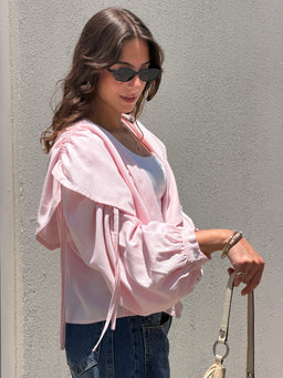 - Product: The Cloud Cropped Shirt. A light, pastel pink cropped hoodie-style jacket layered over a white top. The model is wearing it with dark jeans and sunglasses, giving a casual, chic look.
- Audience: Women (judging by the female model, styling, and cropped silhouette).
- Notable attributes: Soft pink color, lightweight, cropped length with an oversized hood and drawstrings, long billowy sleeves with gathered cuffs, and a relaxed casual style. Material appears to be a lightweight knit or sweatshirt fabric.