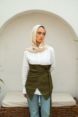 - Product: Willow Corset (front-of-temale styling shown as an adjustable wrap corset over a white shirt)
- Audience: Women
- Notable attributes: olive green wrap-over corset over a white long-sleeve shirt, layered look with a beige hijab/scarf, light blue jeans, and a neutral backdrop with a arched architectural setting. Material appears to be a structured fabric with a wrap tie detail at the waist. Color palette includes olive green, white, beige, and light denim. Style: modest, layered, contemporary wrap corset over blouse.
