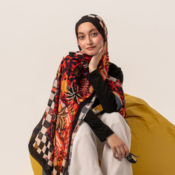 A lightweight chiffon scarf draped over a model, with one end wrapped around the shoulders and the other hanging down. The design features bold, colorful patterns in red, black, and gold hues, with a geometric checkerboard border along the edge. The scarf is styled for a modest, head-and-shoulders look, suggesting a unisex to women's audience, and appears sheer and flowy with a vibrant, tropical-floral motif.