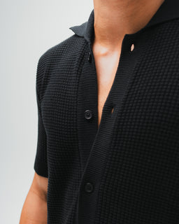 - Product: Bayside Loop Knitted Shirt in black. A short-sleeve knit polo-style shirt with a textured, looped knit pattern and a partially open placket.
- Intended audience: Men (based on fit, neckline, and styling with minimal collar), unisex appearance possible but presented as men’s shirt.
- Notable attributes visible: black color, textured waffle/loop knit, short sleeves, button-front placket with dark buttons, polo collar. Fabric appears medium-weight knit with a slightly relaxed fit.