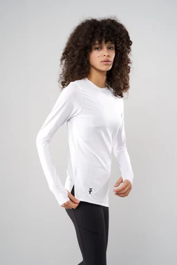 The image shows a white, long-sleeve performance top with a fitted, athletic silhouette. It appears aimed at women, modeled by a female with a lean build; the fabric looks smooth and lightweight with a slight sheen, suggesting moisture-wicking material. Details include a crew neck, clean front with minimal seam lines, and a small black logo near the lower hem. The top is paired with black leggings, reinforcing a sporty, activewear look.