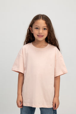 A light pink oversized unisex T-shirt with a relaxed, boxy fit. It’s modeled by a young girl, suggesting a youth-friendly sizing look; short sleeves that reach above the elbow and a classic crew neckline. The fabric appears soft cotton or cotton-blend with a solid plain color and minimal visible detailing, giving a casual, minimalist style.