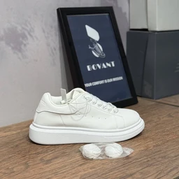 - A white low-top sneaker with a rounded toe and chunky sole sits on a wooden surface. The shoe features subtle stitched panels and small perforations along the side, with matching white laces and a lightly padded collar. A pair of extra laces is placed in front, and a framed display in the background suggests a fashion or design-forward presentation. Intended audience appears unisex or general (adults), based on the neutral styling. Notable attributes: white leather or faux leather upper, thick platform sole, minimalist design, clean and modern look.
