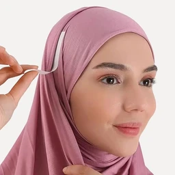 - What the product is: A beauty/soft-hold hijab pin (سبّالة تثبيت الحجاب) being used to secure a hijab.
- Apparent audience: Women (unisex not applicable here; audience inferred as women based on hijab styling).
- Notable attributes: The pin is slender with a small curved end, shown in use to secure a pink hijab. Visible color is light pink; style is simple, minimal, and discreet for hijab fastening. The model wears a matching pink hijab and light makeup for a clean, everyday look.