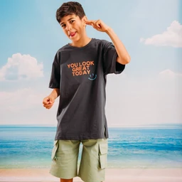 The product is a pair of mint green cargo shorts paired with a dark gray t-shirt featuring a graphic text. The photo shows a young male models wearing the outfit in a casual, sunny beach setting, suggesting a unisex or boys’/men’s casual wear audience based on sizing and styling. Notable attributes include the mint green cargo shorts with side pockets and a dark T-shirt with an orange text graphic and a small smiley, presenting a relaxed, summer-ready look.