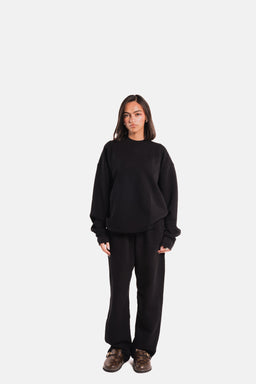 - What the product is: A black heavyweight crewneck sweatshirt paired with matching black sweatpants, shown as a coordinated loungewear set.
- Apparent audience: Unisex or possibly men’s; model appears to be a young adult female, but styling and sizing suggest a relaxed, oversized fit suitable for men or women who prefer a loose cut.
- Notable attributes: Thick, structured black fleece fabric with long sleeves and ribbed cuffs; crewneck collar; relaxed/oversized silhouette; pants with a comfortable waistband and wide leg. No visible branding.