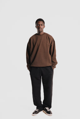 - A brown heavyweight crewneck sweatshirt styled on a male model, paired with black relaxed-fit pants and black-and-white sneakers. The look is casual and minimalist, with a neutral espresso brown top and dark bottoms.
- Visible materials and colors: medium-weight brown fleece/knit fabric for the sweatshirt; black pants; black sneakers with white accents. The fit appears oversized or relaxed, with dropped shoulders and a slightly cropped hem.