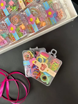 - What the product is: Small clear plastic storage boxes containing a variety of colorful elastic hair ties, mini bracelets, and small accessories. The set appears to be a “Tie Box” collection with multiple compartments for organizing.

- Apparent audience: Unisex or girls’ use, judging from the bright pastel colors, flower-themed packaging, and small, playful accessories suited for children or teen styling.

- Notable attributes: Visible pastel and neon colors (pink, yellow, green, blue, purple), assortment of hair ties and decorative mini items; compact, snap-closable compartments with individual slots; flower-themed packaging with playful cartoon motifs. The boxes are clear, showing the contents, and have small hooks or loops for hanging.