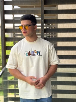 - What the product is: A light-colored, oversized short-sleeve knit tee with an exposed-stitch detail and a bold graphic on the chest reading “IRON” with small images inside the letters.
- Apparent audience: Men’s (model styling, relaxed fit, short sleeves, and neutral oversized look suggest masculine cut).
- Notable attributes: Heavy waffle knit texture, exposed-stitch construction, off-white/cream fabric, oversized/boxy silhouette, graphic print with multicolor letter-inset imagery, and a casual streetwear vibe. The model wears mirrored yellow sunglasses to finish the look.