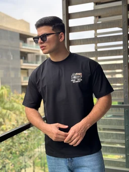 A black minimalist oversized t-shirt worn by a male model. The shirt features a small, white printed graphic on the left chest with simple branding elements. The overall look is casual and modern, with short sleeves and a relaxed fit, paired with light blue jeans. Notable visible attributes: solid black color, lightweight fabric, relaxed oversized cut, and a small white graphic/logo on the chest. Audience inferred: men’s. Material not clearly identifiable from the image, only the fabric appearance suggests a soft jersey knit. Color: black; Style: minimalist oversized tee.