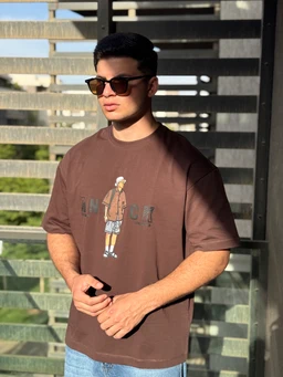 - Product: An oversized short-sleeve tee in brown, featuring a small graphic on the chest of a cartoon-like figure wearing shorts and a jacket.
- Audience, styling, and materials: Appears designed for men (model and fit suggest male presentation) with an oversized cut and relaxed fit. The fabric looks like a standard cotton knit with a matte finish and a vintage acid-wash vibe. 
- Notable attributes: Brown color, graphic print on the front, crew neck, short sleeves, casual streetwear style. No visible branding besides the graphic.