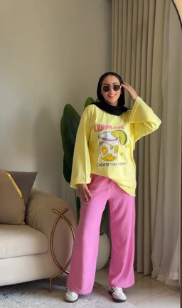 This is a casual graphic T-shirt and wide-leg pants set. The model appears to be a woman, suggesting a women’s/mixed audience look, with a bright yellow oversized long-sleeve T-shirt featuring a printed graphic and pink relaxed-fit pants. Notable attributes include the lemon-themed front print on the shirt, the bright lime-yellow color of the top, the vivid pink pants, and white sneakers with round sunglasses as styling. The overall style is playful, bold color-block casual wear, likely intended for everyday leisure.