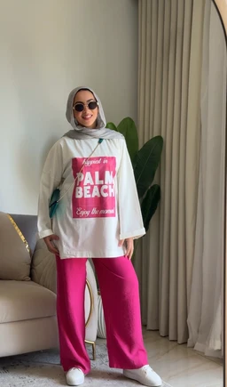 A casual unisex or women’s oversized T-shirt with a bold pink graphic on the front that reads “Palm Beach.” The model wears a light hijab and round sunglasses, paired with bright pink wide-leg pants and white sneakers, suggesting a relaxed, streetwear vibe. Visible materials appear to be a soft cotton knit in off-white, with a large rectangular pink print on the chest and a relaxed, long-sleeve silhouette.