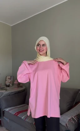 - Description: A pink short-sleeve T-shirt with a loose, oversized fit, worn over a light cream hijab. The model is posing in a casual living room setting, standing and holding the top edges of the shirt.
- Audience attributes: Appears intended for women (feminine styling, hijab pairing), sized as loose/streetwear.
- Visible materials and style: Smooth knit fabric in soft pink; round neckline, relaxed silhouette with dropped shoulders and wide sleeves, casual everyday tee style. Not showing any branding.