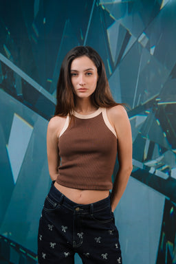 A brown two-tone sleeveless tank top with a light beige neckline and armhole trim, worn by a female model. The model appears to be showing the top as casual, likely for women; styling includes dark jeans with small light-colored embroidery. The visible materials suggest a stretchy knit fabric in brown with beige contrast trim, giving a sporty, everyday look.