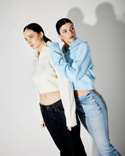 - Product: A half-zip sweater worn by two models. The styling is casual athleisure with cropped silhouettes.
- Audience: Appears unisex or women-focused, judging by the cropped length and fit on the female models.
- Notable attributes: Textured knit fabric in light cream and light blue; high collar with half-zip opening; long sleeves; relaxed yet fitted cropped hem. Models wear with jeans for a casual streetwear look.