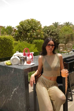 Product is a coordinated two-piece lounge set (crop top and high-waisted leggings) in a shimmery champagne-beige color. The target audience appears to be women, judging by the sleeveless cropped top and fitted leggings. Notable features: metallic/sparkly knit fabric, sleeveless halter-like cropped top, matching high-waisted pants, soft and stretchy texture, laid-back poolside styling with a beverage and outdoor garden backdrop. No visible branding.