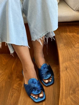 - What the product is: A pair of metallic wrap-around sandals with large decorative blue bows on the straps.
- Apparent audience: Women, judging by the foot-shape, open-toe style, and styling with cropped frayed jeans.
- Notable attributes: Midnight blue metallic straps, wide woven bow detail, open-toe design, square/rounded toe, flat sole with a textured footbed. The overall look is casual, footwear for warm weather.