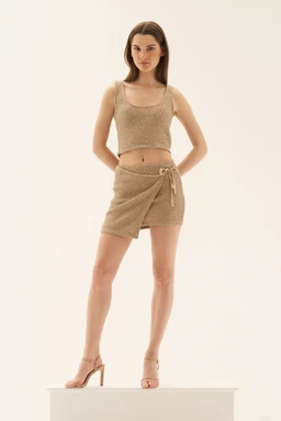 - Description: A matching two-piece set consisting of a sleeveless crop top and wrap-front shorts in a metallic gold-beige knit. The fabric has a subtle shimmer and a textured knit look; the top is cropped, the shorts sit mid-thigh with a tied belt detail at the waist. The model is wearing open-toe high-heeled sandals.

- Audience and styling cues: Appears styled for a women’s market, with a sleek, fitted silhouette and minimal accessories, suitable for going out or casual glam looks. Notable attributes: gold-toned shimmer knit fabric, crop top, wrap shorts with side tie, sleeveless tank top, neutral metallic color.