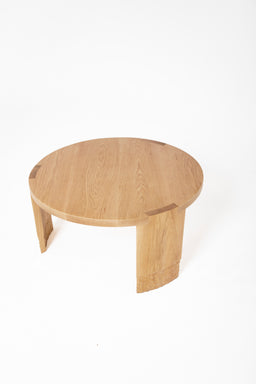 - A round wooden coffee table with a light oak finish. It has a smooth, flat top and two wide, curved legs that join the underside to form a sturdy base. The visible grain and color suggest a natural wood material with a simple, minimalist style suitable for modern interiors. Target audience: unisex. Notable attributes: round top, light wood tone, clean lines, two chunky side supports.