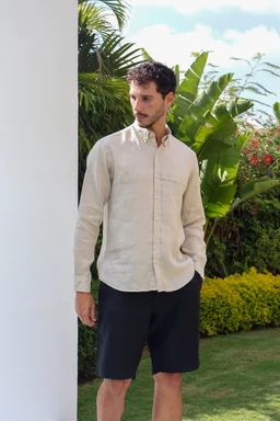 - What the product is: A beige premium linen shirt worn by a male model.
- Apparent audience: Men (adult/masculine styling), suitable for casual to smart-casual wear.
- Notable attributes visible: Light beige linen fabric with a relaxed, button-front shirt design, long sleeves (buttoned cuffs), collared neckline, and a classic chest pocket. The model pairs it with dark shorts and a tropical outdoor backdrop.