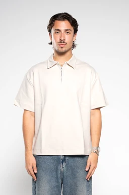 - A light beige zip-up polo with short sleeves and a polo collar, worn by a male model. The shirt has a relaxed, boxy fit and a short zip placket at the neckline.
- Intended audience: men's.
- Notable attributes: earth/beige color, short-sleeve polo style, zip front, mid-hip length, plain knit fabric with minimal detailing.