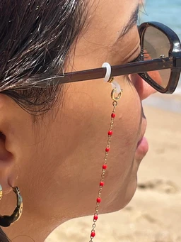 This is a sun/eyeglass chain featuring a delicate beaded strand that drapes from the ear to the chest. It appears designed for women's wear, attached to the temple with a small ring and secured by a white loop over the ear piece. The chain has small red beads linked along a fine gold-tone chain, with a short dangling drop and hoop accents, giving a light, tropical accessory vibe.