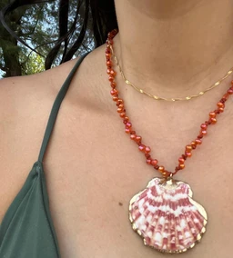 - What the product is: A seashell pendant necklace set on two contrasting cords. The focal piece is a pink and white scallop shell framed in a gold-tone edge, hanging from beaded coral-colored and gold-toned chains.
- Intended audience: Women's jewelry based on the styling, draped over a female neckline in a halter-style top.
- Notable materials, color, and style: Pink and white patterned scallop shell pendant with a gold-tone rim, paired with coral-toned bead strand and a delicate gold-toned chain. Overall style is boho/bohemian coastal.