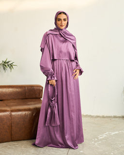 - Product: A lilac satin maxi dress with matching hijab, model wearing a full-coverage look.
- Intended audience: Women (modest, with hijab styling), unisex-adjacent modest fashion.
- Visible attributes: Soft lilac satin fabric with a loose, flowing silhouette, long puff sleeves with gathered cuffs, high neckline with draped scarf detail, floor-length skirt, matching headscarf draped over the shoulders. The material appears smooth and shiny satin with a slightly structured bodice and a gathered waist. No brand markings visible.