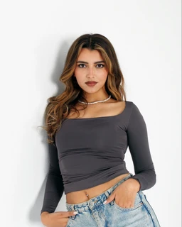 This is a Curve Fit Top in Graphite, shown on a female model. The top has a fitted, long-sleeve, off-the-shoulder/boat neckline silhouette with a cropped length. Visible attributes include a solid graphite/charcoal color, stretchy jersey fabric, and a form-fitting, square-cut neckline that sits off the shoulders. The model pairs it with light-wash jeans and a simple choker, suggesting a casual-to-chic styling.