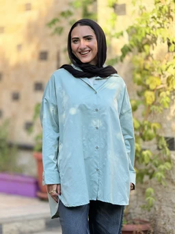A light blue poplin shirt with a loose, relaxed fit and a button-front placket. The model appears to be a woman, dressed modestly with a dark headscarf, long sleeves, and an oversized silhouette suitable for casual or everyday wear. Key visible features include the soft sheen of poplin fabric, drop-shoulder seams, and a longer length that drapes over the hips.