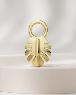 A gold-toned charm in the shape of a stylized leaf or monstera leaf, with a circular loop at the top for threading onto a bracelet or necklace. Appears unisex in style, leaning toward a chic, tropical-inspired accessory. The surface is polished, reflecting light to emphasize the smooth, metallic finish. No visible branding.