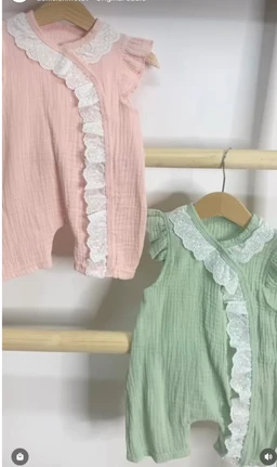 This is a romper-style baby or toddler one-piece outfit, shown on small hangers. It appears unisex with delicate ruffled white lace trim along the front placket and collar, in soft pastel pink and mint green. The fabric looks lightweight and crinkled, with short sleeves and a loose, comfortable fit suitable for warm weather.