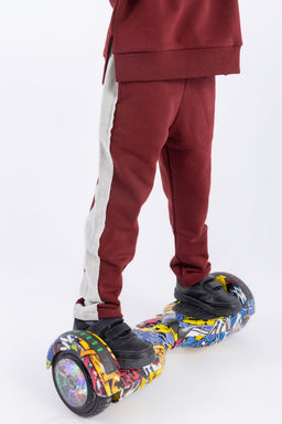 - What the product is: A pair of jogger-style pants with a two-tone design, shown worn by a model on a hoverboard.
- Apparent audience: Unisex or boys/men based on fit and styling; the model’s build and the streetwear vibe suggest a youth-to-adult casual market.
- Notable attributes: Deep burgundy fabric with a white side stripe running down the leg. The material appears to be a soft knit or fleece. The look is sporty and relaxed, with a tapered ankle cuff. The photo emphasizes the color contrast and a casual athleisure style.